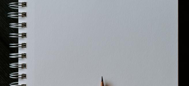 The blank page of a spiral-bound sketchbook page. The sharpened point of a pencil waits at the edge of the page.