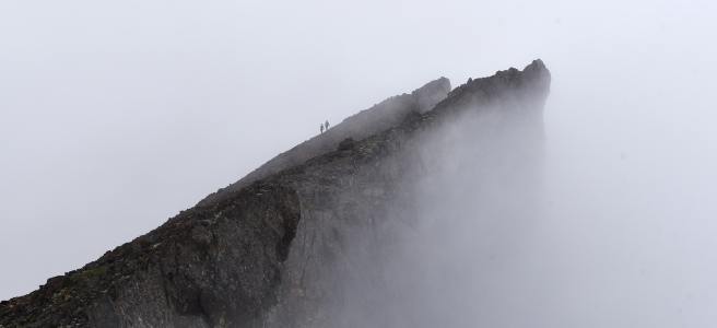 Two tiny figures in silhouette against the fog. They stand on top of a jagged spur of black rock.